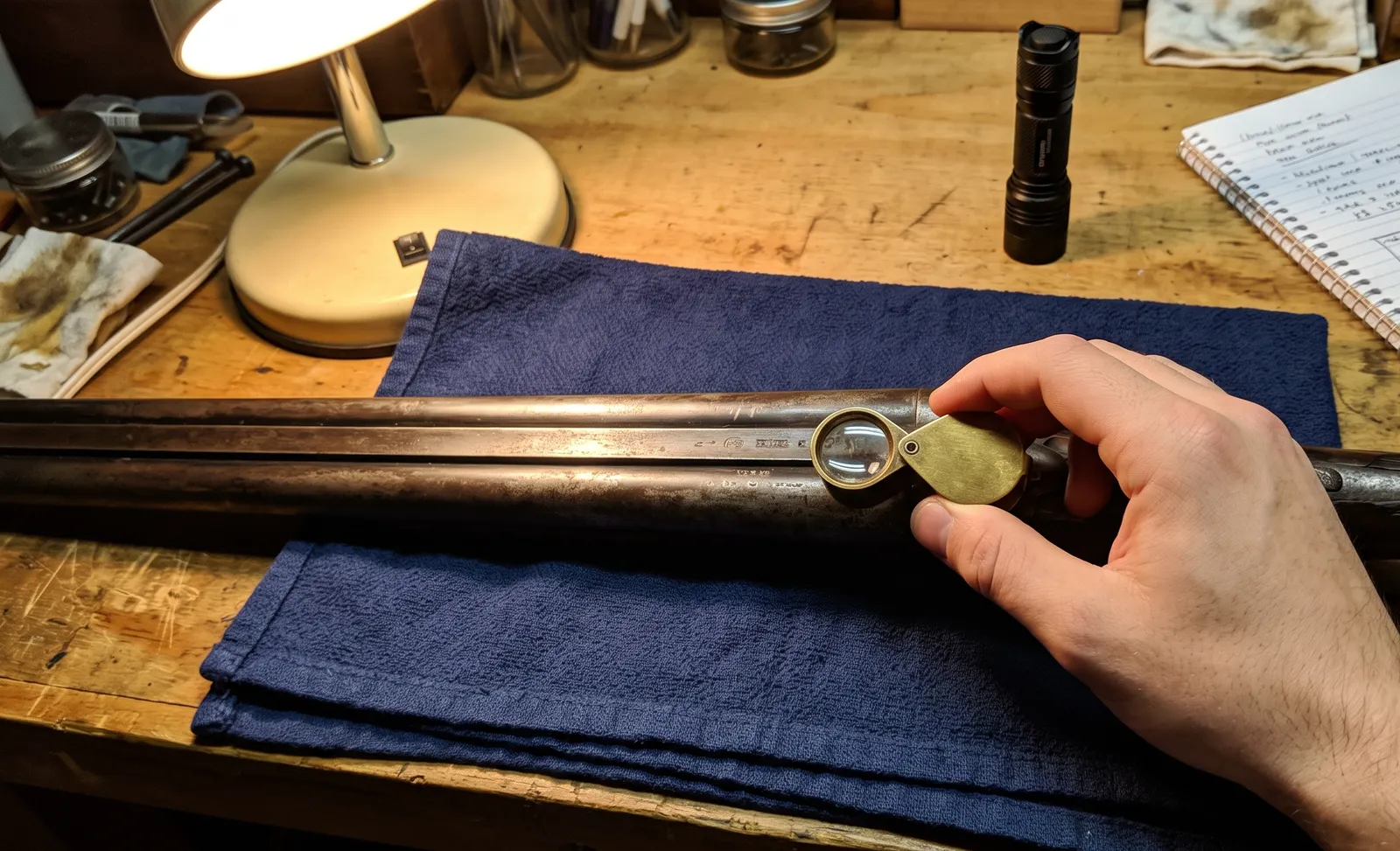 Examen à la loupe du dessous des canons d'un fusil juxtaposé posé sur une serviette, lampe et carnet à côté